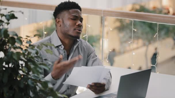 Konsantre olmuş sinirli Afro-Amerikan adam dizüstü bilgisayarla oturmuş evraklara bakıyor bankadan kötü haber gelen kağıt bildirimi okuyor ve kendini üzgün hissediyor. — Stok video