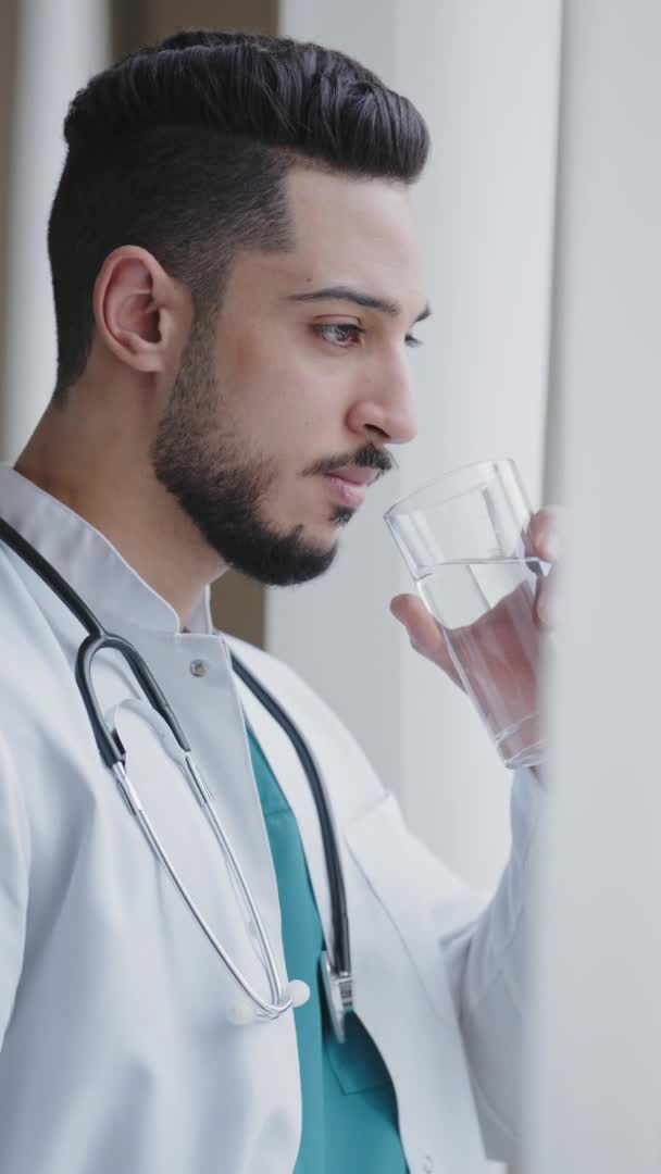 Vertical view of young man thoughtful arabian doctor wear white medical coat looking through window drinking water serious sad medical worker think about ending quarantine in future healthcare concept — Stock Video