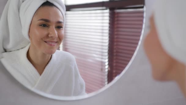 수건을 머리에 쓰고 목욕 한 젊은 여성 이 거울을 닦은 후 얼굴은 건강하고 부드럽고 습기가 많은 피부 화장품 제거 화장은 자연 미용 스킨케어 치료 개념을 즐긴다 — 비디오