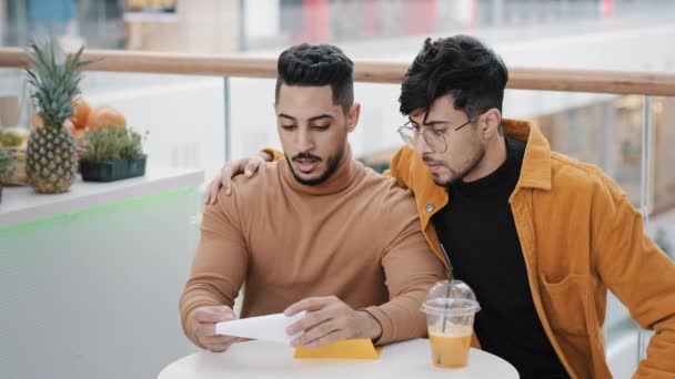 Two friends students sitting in cafe young worried man receiving letter reading bad news upset feels sad failing exam dismissal financial problem loss arab guy comforting best friend hugging cheers up — ストック動画