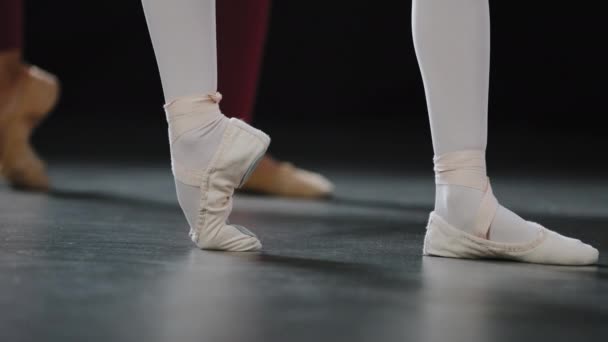 Две пары женских ног на полу на сцене перформанса в студии танцевального класса выполняют балетные упражнения, растягивающие ноги, стоят на пуантах, балерины танцовщицы повторяют движения учителя-тренера — стоковое видео