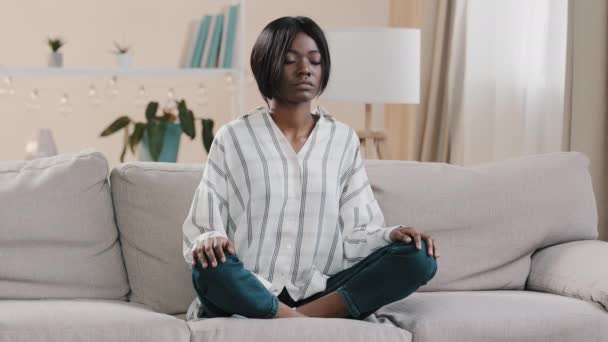 Young focused african american woman sitting in lotus position with closed eyes healthy calm girl relaxing on sofa calming down taking deep breath meditating relaxing at home getting rid of stress — Stock Video