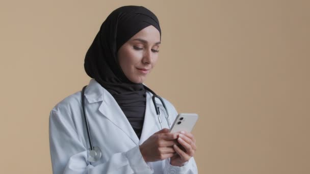 Artsen vrouwelijke arts therapeut vrouw dragen hijab wit uniform staan in de kliniek met behulp van smartphone mobiele gezondheidszorg toepassing arabisch medisch chatten consulting patiënten online telegeneeskunde concept — Stockvideo