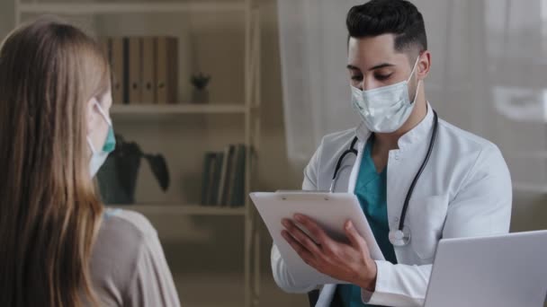 Arabische mannelijke arts arts in medisch gezichtsmasker het maken van notities op klembord consulting vrouwelijke patiënt uit te leggen medische resultaten geven recept behandeling benoemen overleg zitten in kliniek — Stockvideo