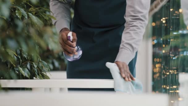 Önlüklü Afro-Amerikan erkek garson restoranın açılışından önce dezenfektanla dezenfekte masasını temizliyor. — Stok video