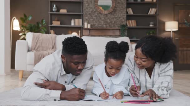 Famille africaine parents mère et père avec fille enfant enfant dessin image à colorier avec des crayons couchés sur le sol à la maison maman et papa aider écolière avec l'art thérapie profiter de jouer dessiner ensemble — Video