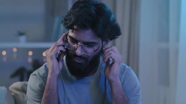 Arabic Indian bearded man with glasses talking on phone at house party sitting on sofa neon light hard to hear conversation loud music noisy birthday celebration answering phone call trying listening — Stock Video