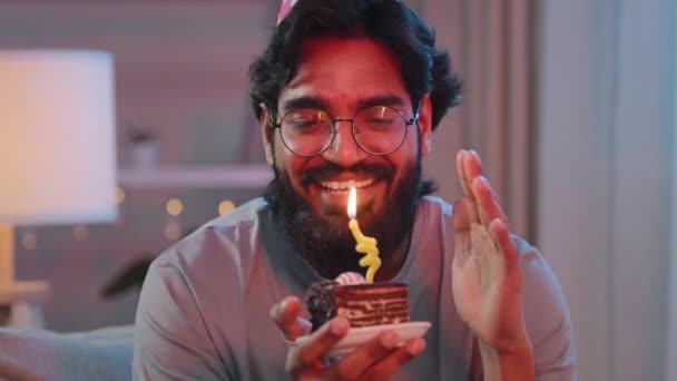Retrato árabe feliz indio hombre barbudo cara lleva festivo cumpleaños rosa sombrero y gafas sostiene cupcake hace deseo alegre felicitación en casa soplando velas ardientes en pastel fiesta de celebración — Vídeos de Stock