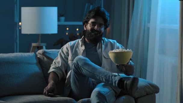 Arab adult bearded man in glasses indian male bachelor after work sits on sofa in dark living room turns on TV with remote control choosing channel watching online movie film program eating popcorn — Stock Video