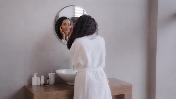 Молодая счастливая женщина, стоящая в ванной комнате, глядя в зеркало трогательное лицо после нанесения крема наслаждается здоровой увлажненной кожей, любуясь танцами, наслаждаясь энергичной утренней гигиенической рутиной — стоковое видео
