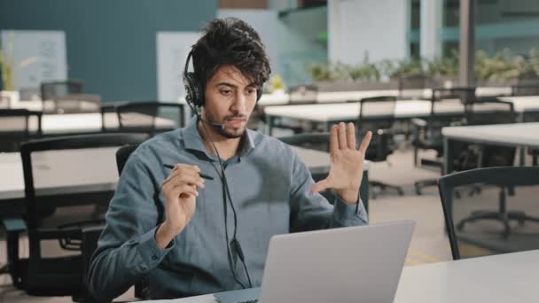 若いインドのビジネスマンの投資家は、オフィスで従業員遠くのコンサルティングクライアント仮想トレーニングコンセプトとノートパソコンの交渉トークオンラインでビデオ通話会議でヘッドフォン通信を身に着けている — ストック動画