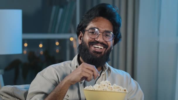 Πορτρέτο αραβικός ισπανόφωνος Λατίνος Ινδός μουσουλμάνος γενειοφόρος άνθρωπος χαρούμενος χαλαρός τύπος σε γυαλιά βλέποντας ταινία πρόγραμμα αστεία σειρά τηλεόραση online τρώει ποπ κορν γέλιο διασκεδάζοντας στο σπίτι το βράδυ σκοτάδι — Αρχείο Βίντεο