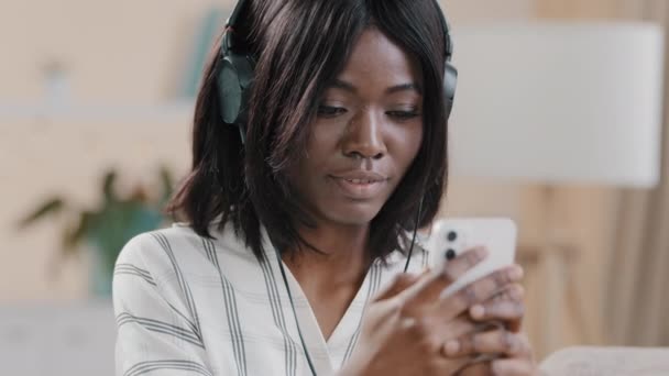 Close-up happy young pretty African american woman holding smartphone słuchanie przyjemnej muzyki na słuchawki taniec słucha utworu ulubiona piosenka audio czuje się zadowolony korzystając weekend hobby i odpoczynku — Wideo stockowe
