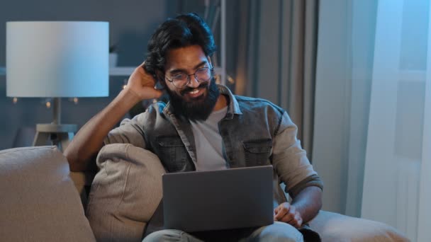 夜深时分，阿拉伯印第安人大胡子男子千禧年商人自由职业者坐在家里的沙发上，用笔记本电脑远程交谈，在网上摄像头的视频电话会议上挥手致意 — 图库视频影像