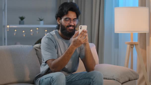 拿着现代智能手机发短信的快乐的阿拉伯印度男性千年男人，戴着眼镜坐在沙发上，用手机应用程序观看网上搞笑视频 — 图库视频影像