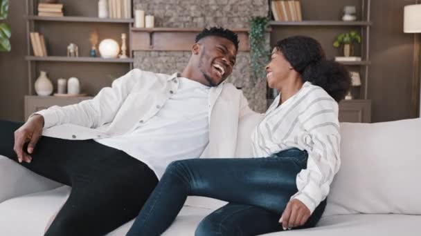 幸せなアフリカ系アメリカ人のカップルジャンプは柔らかいソファの上に落ちる快適なソファ笑いは、自宅を借りフラットへの移動日を祝う新しい家具の不動産移転の残りの部分をリラックスお楽しみください。住宅所有者の家族 — ストック動画