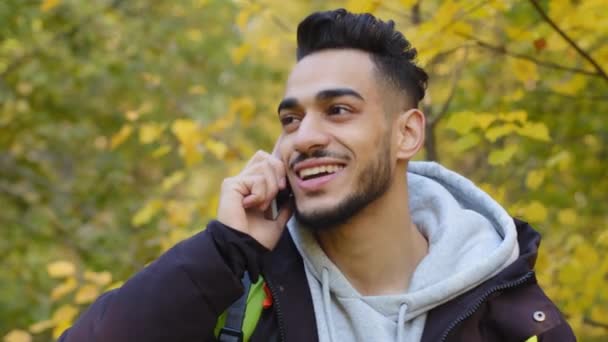 Close-up szczęśliwy młody, przystojny arabski mężczyzna stojący na świeżym powietrzu turysta podróżuje w przyrodzie uśmiechając się rozmawiając przez telefon komórkowy dzieląc się dobrym doświadczeniem komunikowania się z przyjacielem za pomocą smartfona komunikuje się zdalnie — Wideo stockowe