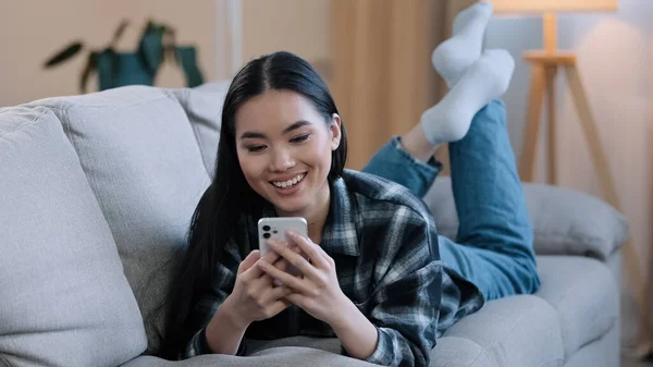 Aziatisch vrouw ontspannen meisje vrouw liggend op de bank thuis genieten chatten typen app mobiel telefoon ontspannen kijken viraal live video spelen spel volgen als post in social media delen en stuur glimlach emoji scroll web — Stockfoto