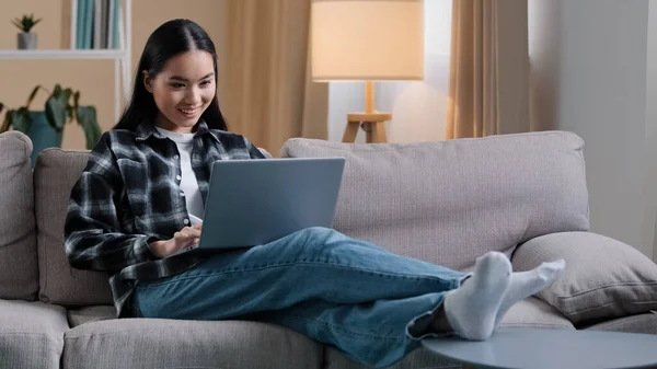 Carefree Asian girl enters living room sit down on comfortable sofa takes laptop with smile and watches video online woman uses computer sitting on couch at home casual browsing net resting typing — Photo