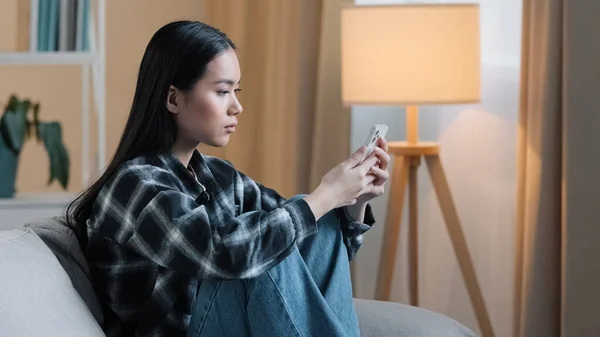 Vista lateral de triste aburrido mujer asiática chica sentada en el sofá en casa con el teléfono de la señora infeliz grave desplazamiento de navegación en el teléfono inteligente y el uso de la aplicación de teléfono móvil viendo noticias de redes sociales alimentación mecanografía — Foto de Stock