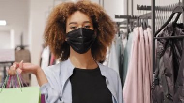 Kapalı gişe Afrikalı kadın tüketici Kıvırcık kadın müşteri yüz koruma maskeli müşteri giyim mağazasında alışveriş poşeti ve salgın hastalık alışverişi yapıyor.