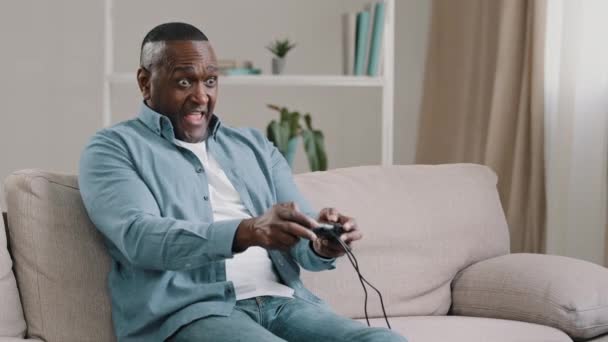 Heyecanlı duygusal yetişkin Afrikalı Amerikalı erkek oyuncu oturma odasında oturmuş video oyunu oynuyor kumandayı elinde tutarak bilgisayar oyunları oynuyor konsol oyunu oynuyor. — Stok video