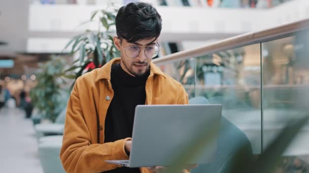 Young indian man looking at laptop screen checking email smiling enjoying free time browsing web page on dating site making order in online store chatting on social media using computer application — ストック動画