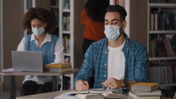 Aşırı yorgun, koruyucu maskeli genç Arap erkek öğrenci kütüphanede oturuyor ödev hazırlıyor sınava hazırlanıyor, hata yapıyor elle başını kavrıyor şok krizi geçiriyor. — Stok video