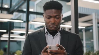 Ciddi Afro-Amerikan ofis çalışanı akıllı telefon internetini araştırıyor. Arkadaşlarıyla çevrimiçi sohbet etmeyi seçiyor. Düşünceli erkek kullanıcı tarayıcı hesabına tıklıyor.