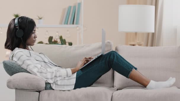 Młoda studentka freelancer leżąca na kanapie w domu, pisząca na laptopie słuchająca muzyki na słuchawkach, ciesząca się przyjemną piosenką, odpoczywająca, relaksująca, sprawdzająca pocztę elektroniczną, przeglądająca portale społecznościowe, pracująca zdalnie — Wideo stockowe
