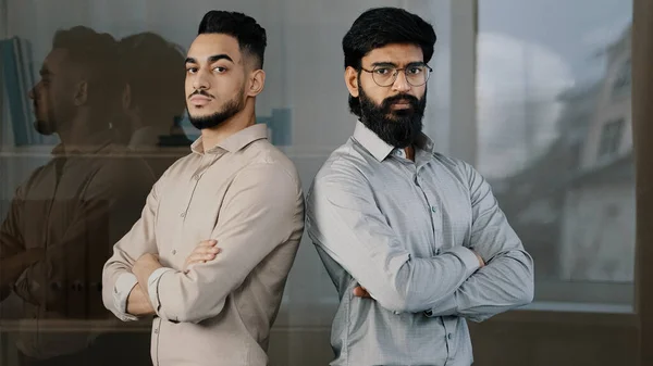 Multiracial couple diverse businesspeople colleagues male co-workers partners arabian bearded man in glasses hispanic young guy standing indoors looking at camera with serious expression cross arms — 스톡 사진