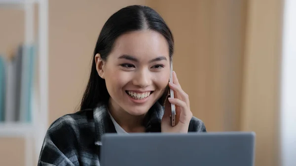 Happy smiling Asian girl Korean female freelancer chatting with friends on phone answering call friendly looking at laptop screen remote working business negotiation making online order by smartphone — стоковое фото