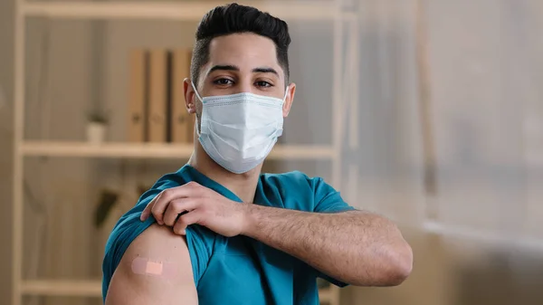 Young arabian man practitioner doctor nurse stand indoor in protective surgical face mask demonstrate adhesive bandage on shoulder after vaccine injection stop covid-19 pandemic coronavirus infection — Foto Stock