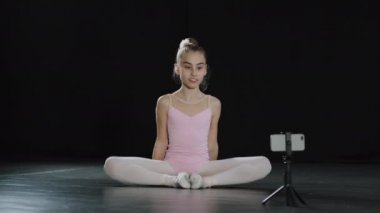 Genç dansçı kız jimnastikçi akrobat balerin çocuk kelebek esneme egzersizi yapıyor dans stüdyosunun kalçaları üzerinde oturmuş internetten uzaktan kumandalı interneti arıyor üç ayaklı telefonuyla.