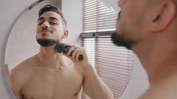 Visão traseira por trás da reflexão masculina no espelho no banheiro cara nu barbudo indiano homem árabe morena usa cortador elétrico de barbear para raspar o cabelo de corte do queixo fácil de barbear em casa — Vídeo de Stock
