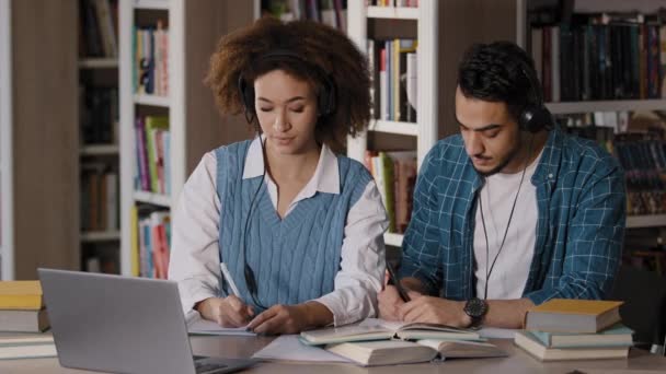 Two diverse students young man and woman sitting at desk listening attentively to teacher lecture at distance on laptop using headphones write notes read textbook study remote team project in college — 비디오