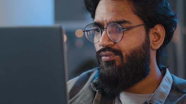 Großaufnahme bärtigen männlichen Gesichts Reflexionslicht von Laptop-Monitor in einer Brille. Beschäftigter, ernsthaft fokussierter arabischer Inder, der abends freiberuflich am Computer arbeitet und nachts im Netz surft — Stockvideo
