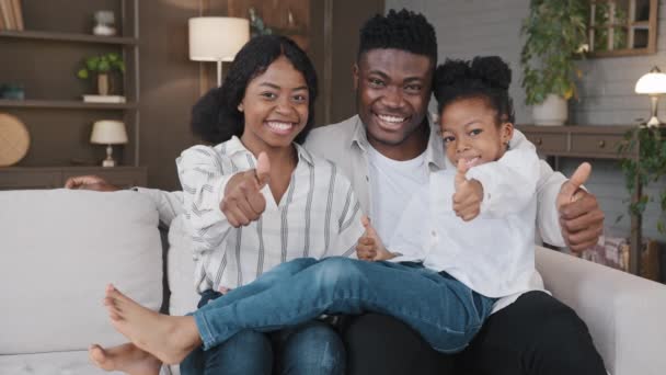 Happy African familie unge værge forældre og lille datter adopteret pige barn sidder på sofaen derhjemme egen lejlighed nyt hus ser på kamera show tommelfingre op godkende anbefale positiv holdning – Stock-video