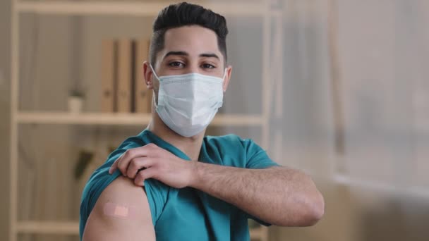 Молодой арабский мужчина практикующий врач медсестра стоять в помещении в защитной хирургической маске лица продемонстрировать клей бинт на плече после инъекции вакцины остановить ковид-19 пандемии коронавируса инфекции — стоковое видео