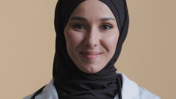 Leende muslimsk arabisk kvinnlig läkare kirurg i hijab traditionell slöja bära medicinsk rock tittar fram på kameran stående poserar i sjukhus positiv allmänläkare närbild ansikte porträtt — Stockvideo