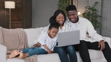 Mutlu Afrikalı aile ve şirin kız çocuğu bilgisayar ekranına bakıp eğlenceli sosyal medya videoları izleyerek evdeki kanepede dinlenirken çevrimiçi video konferansı izleyerek eğleniyorlar.