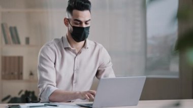 İspanyol Arap erkek iş adamı, koronavirüs karantinası sırasında yazılım uygulaması sohbeti kullanarak sanal ortamda online bilgi ararken dizüstü bilgisayarda yazan maskeli işçi programcısı.