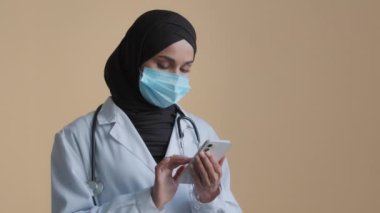 Müslüman maskeli doktor kadın terapist cep telefonlu tesettür beyazı giyiyor. Akıllı telefon tıbbi uygulama. Uzaktan tavsiye mesajı yolluyor.