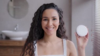 Yakın plan, çekici, mutlu İspanyol bir kadın banyoda kameraya bakıp gülümsüyor, cilt bakım ürünlerinin reklamını yapıyor kavanoza işaret ediyor nemlendirici krem, kanıtlanmış cilt bakımı konseptini destekliyor.