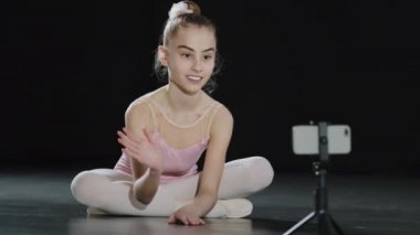 Kafkasyalı kız çocuk balerin jimnastikçi dans salonunda oturmuş cep telefonu kamerasına el sallıyor uzaktan kumandalı öğretmeniyle tripodda akıllı telefon kullanıyor.