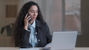 Ciddi İspanyol iş kadını profesyonel girişimci patron ofisteki cep telefonuyla çoklu görev konuşması dizüstü bilgisayar ekranına bak evrak işlerini uzaktan kontrol et internet sitesini onayla