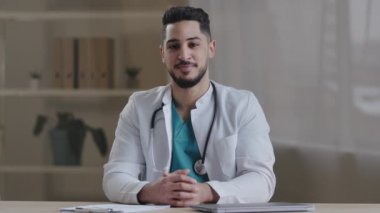 Mutlu gülümseyen İspanyol hemşire doktor kardiyolog stajyer doktor masasında oturmuş kameraların sağlık hizmetlerine bakarak beyaz önlük giyiyor.
