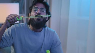 Sakallı, orta yaşlı, komik, Hintli, İspanyol, mutlu, doğum günü çocuğu pembe parti şapkalı, köpüklü sabun köpüğü üfleyen, akşam kutlamalarında evde eğlenen.