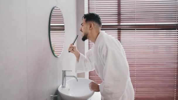 Πλευρά άποψη ξέγνοιαστα αστεία αραβικά ισπανόφωνος ινδός άνθρωπος γενειοφόρος τύπος σε λευκό μπουρνούζι στέκεται στο σπίτι στο μπάνιο κοιτάζοντας αντανάκλαση στον καθρέφτη τραγούδι τραγούδι τραγούδι σε βούρτσα διασκεδάζοντας το πρωί τραγούδι — Αρχείο Βίντεο