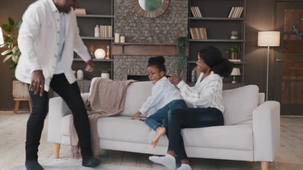 Afrikai család szülei és kislánya gyermek lány ugrás esik kényelmes puha kanapé nevetés szórakozás együtt otthon örül vásárol új otthon költözik a saját lakás játék játék — Stock videók